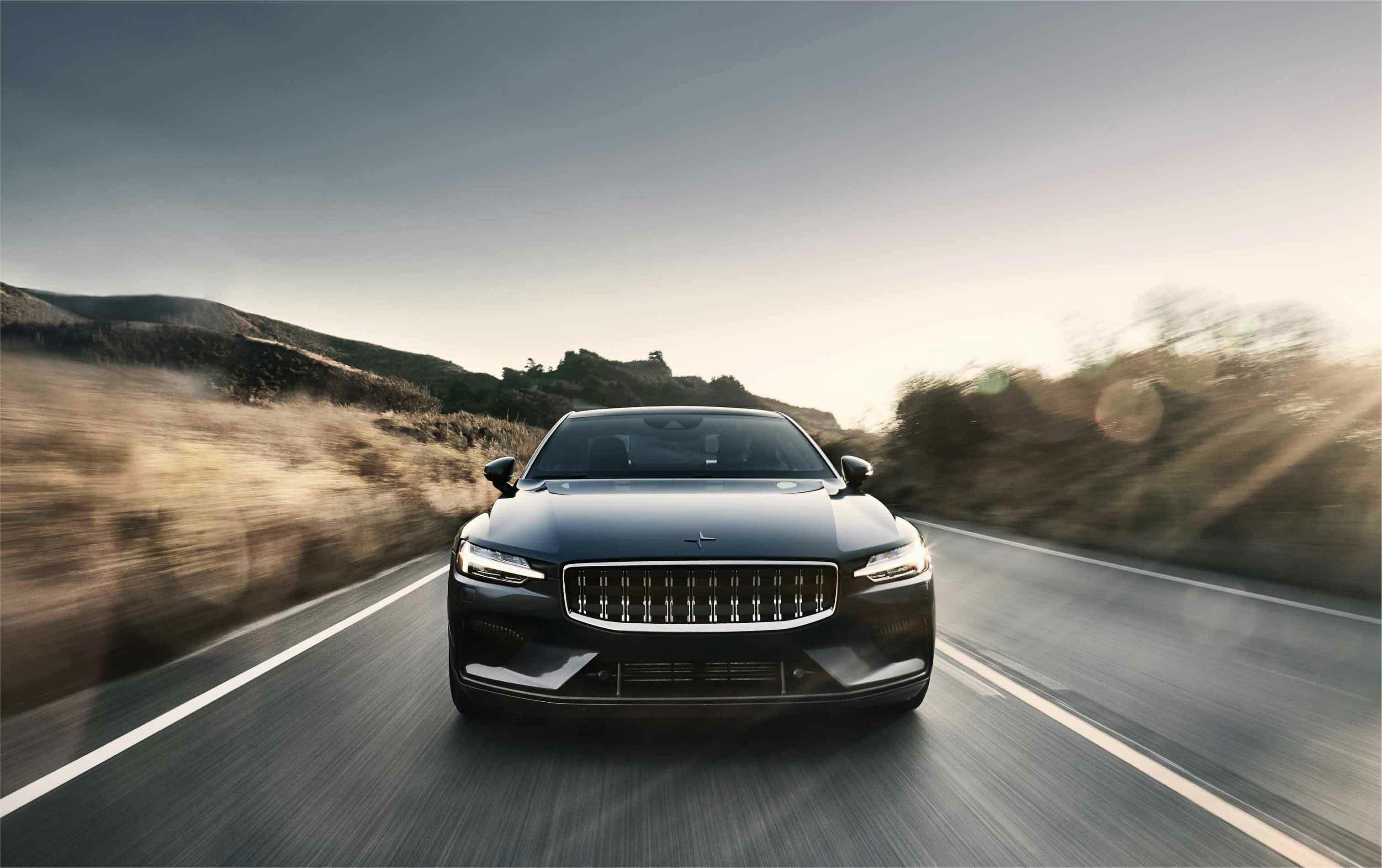 A Polestar black car is driving on an open road with hills in the background, captured from a frontal perspective at sunset